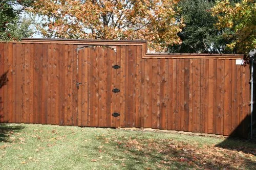 A wooden fence with a gate in a backyard, with trees in the background and grass in the foreground.