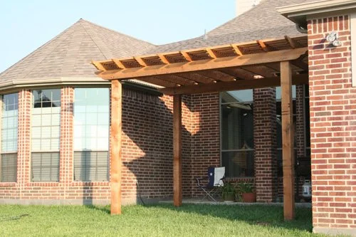 Backyard patio with a wooden pergola attached to a brick house, grass lawn underneath, and a glass door leading inside.