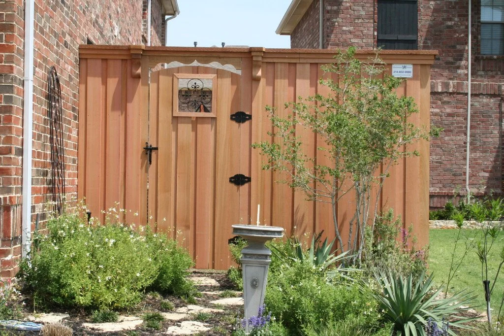 A wooden backyard fence with a small gate, next to a brick house wall, surrounded by plants and bushes, with a birdbath in front.