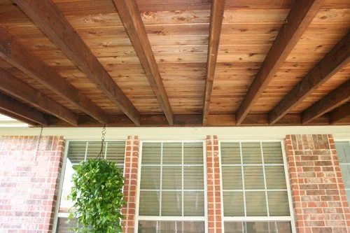 Covered porch with wooden ceiling and a hanging plant next to brick walls and windows.