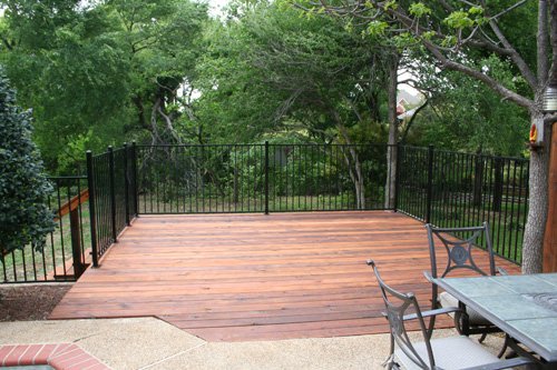 A backyard deck with wooden flooring, surrounded by black metal fencing, with chairs and a table, and lush green trees in the background.