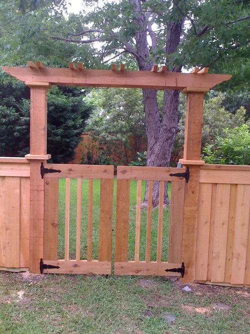A wooden garden gate with a small pergola on top, situated in a backyard with a wooden fence, trees, and grass.