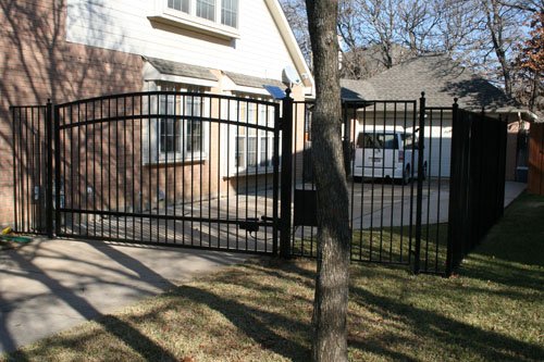 A black metal fence enclosing a front yard with grass, a tree, and a driveway with a white car parked.