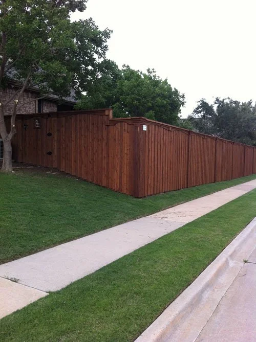 A tall wooden fence along a sidewalk with a grassy lawn in front and trees in the background.