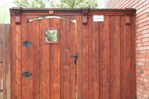 A wooden gate with a small square window, attached to a brick wall on the right, and a small white sign on top.