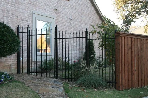 A black metal fence with vertical bars surrounds a small garden area next to a brick house with a window. There is a wooden fence on the right side.