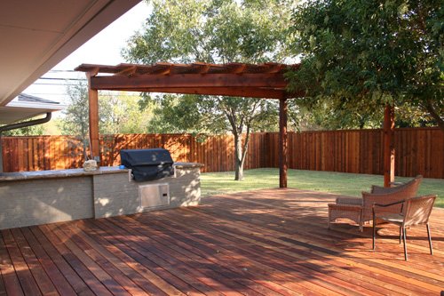 Backyard patio with wooden deck, outdoor seating, a grill, and a wooden fence with trees in the background.