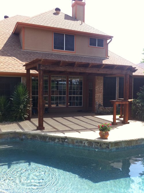 Backyard view of a house with a wooden pergola over a patio, a flower pot, and a swimming pool in the foreground.