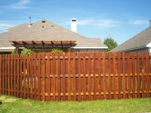 Backyard with a wooden fence, house with a chimney, and a clear blue sky.
