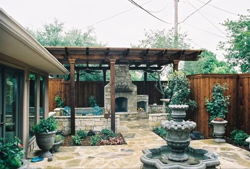 Backyard patio with a stone fireplace, three-tiered fountain, and potted plants, enclosed by a wooden fence.