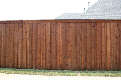 A tall wooden privacy fence along a sidewalk with a house roof visible in the background.