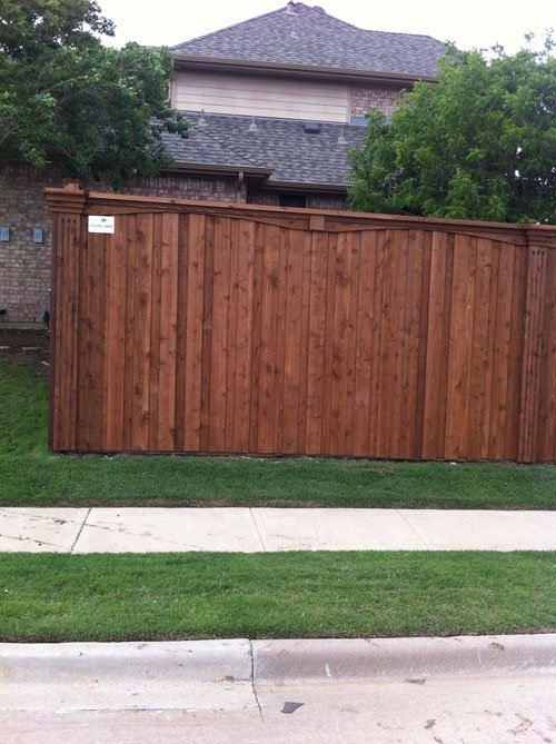 A wooden privacy fence with a gate, a concrete sidewalk, green grass, and a house with a gray roof and trees in the background.
