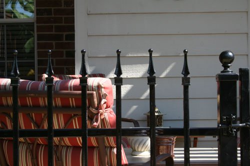 Patio with striped red and beige cushions behind a black metal fence, with a white wooden wall and window in the background.