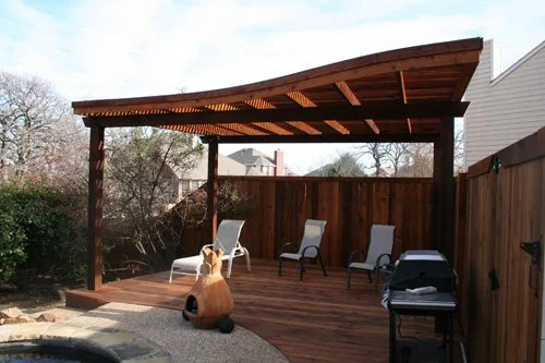Wooden backyard patio with a roof, four chairs, a grill, and decorative pottery, surrounded by trees and fencing.