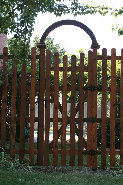 A wooden garden gate with an arch at the top, surrounded by greenery and a backyard with a pool in the background.
