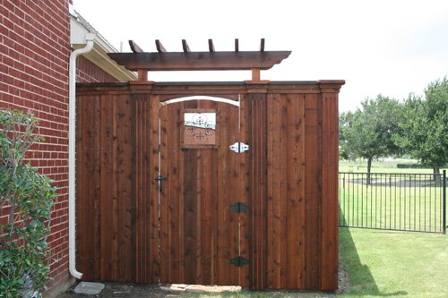 A wooden gate with a decorative top, attached to a brick house wall, with a yard and field in the background.