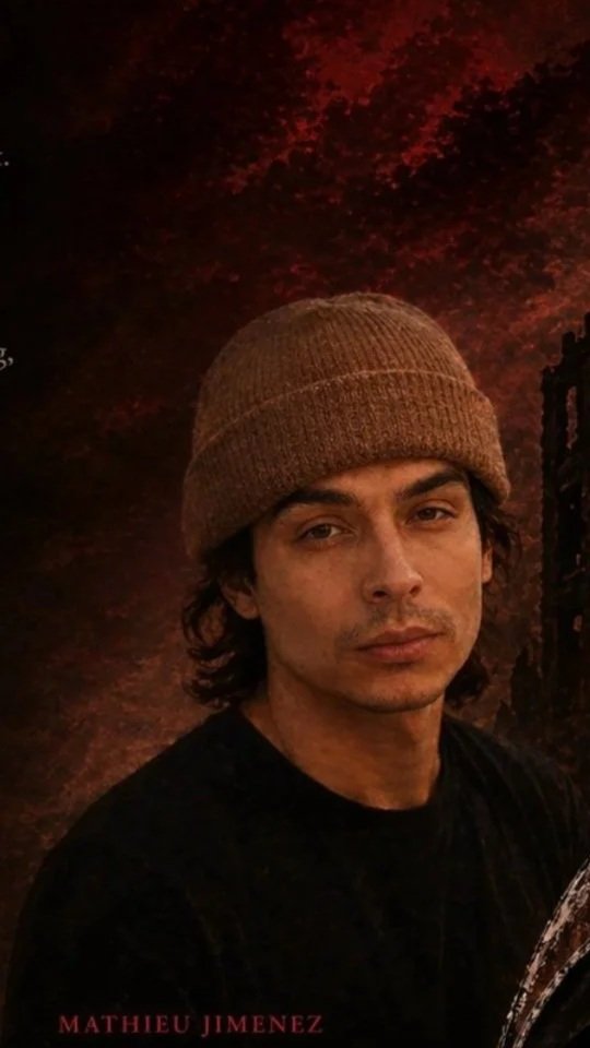 A young man with dark hair, wearing a brown knit beanie, stands outdoors at dusk or night with a dark background featuring red and black foliage.