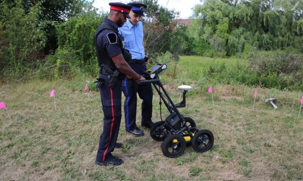 Law enforcement personnel using a Radiodetection LMX100 ground penetrating radar cart to survey an outdoor field.