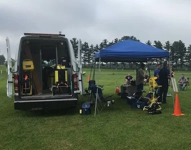 Utility service van with open rear doors and equipment set up beside a canopy tent.