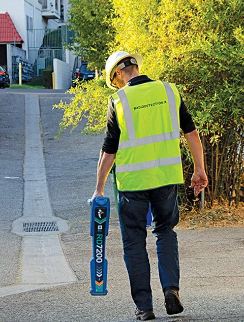 Technician in a hard hat and safety vest walking down a sidewalk while carrying a Radiodetection RD7200 line locator.