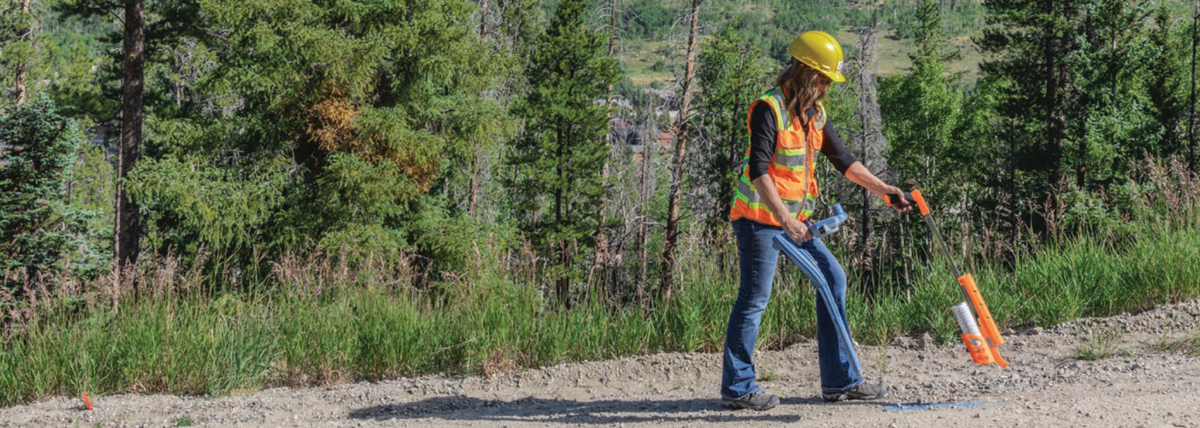 Technician in a hard hat and safety vest using a utility locating transmitter on a rural gravel road.