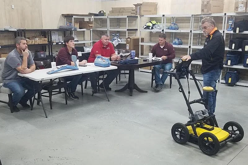 Instructor demonstrating a ground penetrating radar cart to a group seated at tables during a classroom training session.