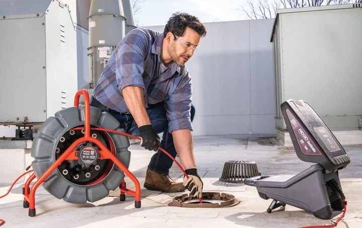 Technician connecting a RIDGID drain inspection camera reel and monitor system to a floor drain outside a building.
