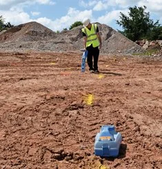 Technician in safety vest performing a utility locating survey with a handheld transmitter across a dirt jobsite.