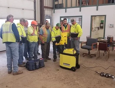 Group of technicians in high-visibility vests gathered indoors for equipment training, with a yellow rolling case.