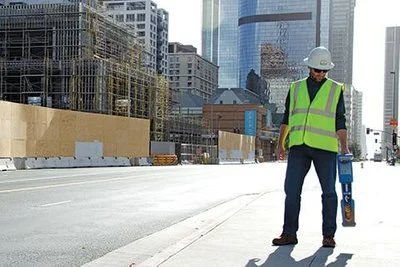 Technician in a hard hat and safety vest holding a Radiodetection RD8200 locator near a downtown construction site.