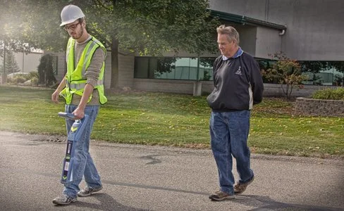 Two men walking together on a residential street, one in a high-visibility vest and hard hat.