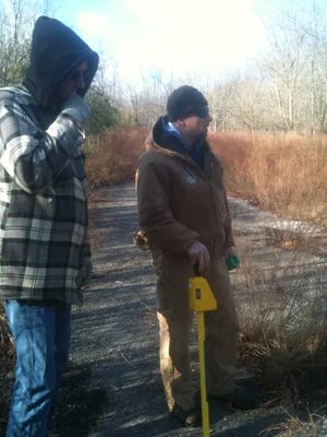 Two technicians outdoors in cold-weather clothing, one holding a locating probe.