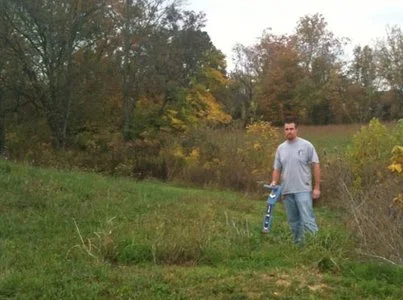 Technician walking a grassy field edge holding a locating device, with autumn foliage in the background.