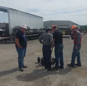 Group of workers in hard hats gathered around utility locating equipment during outdoor training at an industrial yard.