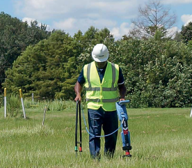 Technician in a hard hat and safety vest operating a utility locator in an open grassy field.
