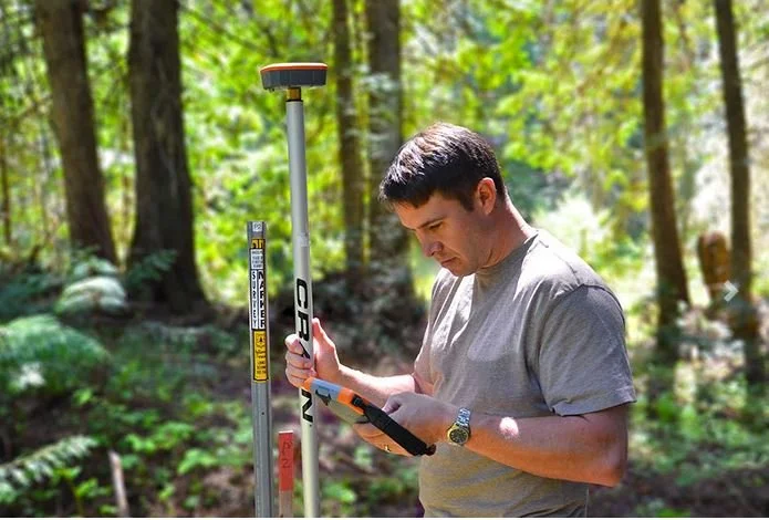 Technician using a GNSS GPS rover pole and handheld controller for field data collection in a wooded area.