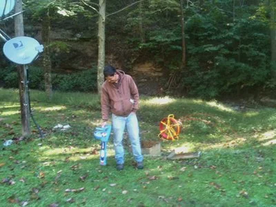 Technician using locating equipment near a blue utility marker sign among trees.