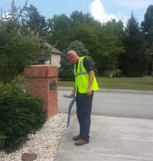 Technician in a high-visibility vest locating utilities near a brick entrance pillar on a gravel driveway.