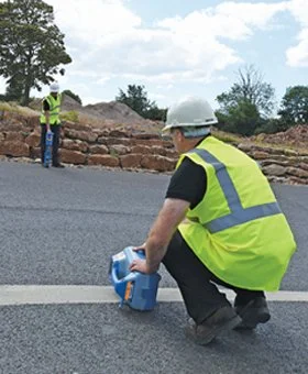 Technician in hard hat and safety vest placing a utility locating transmitter on a paved roadway during a survey.