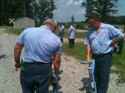 Two technicians using locator wands along a gravel path with a field crew in the background.