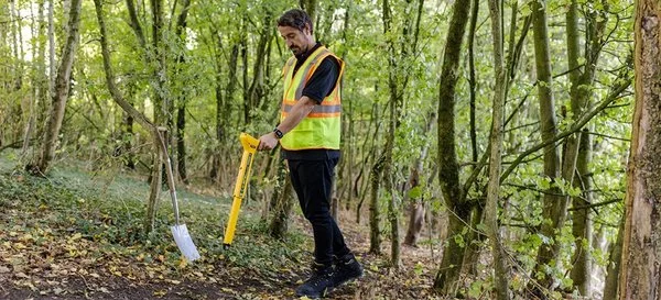 Technician in a safety vest using a handheld utility locator to survey wooded terrain.