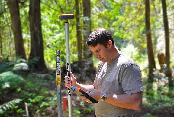 Technician using a GNSS GPS rover pole and handheld controller for field data collection in a wooded area.