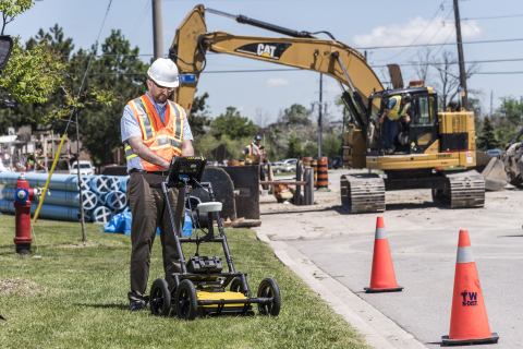 Technician using a ground penetrating radar cart near an excavator at a construction site utility locate.