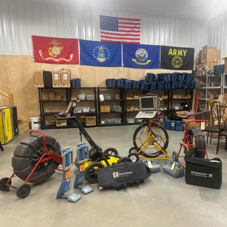 Warehouse display of pipe locating and inspection equipment beneath military branch and American flags.