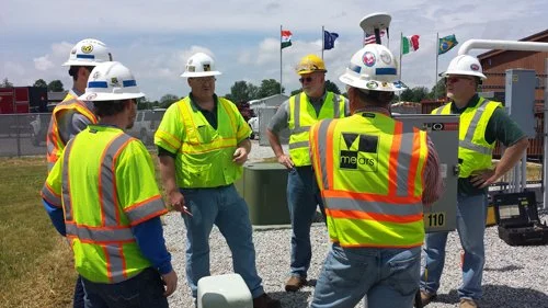 Crew in hard hats and high-visibility vests gathered around utility marking flags during an outdoor training session.