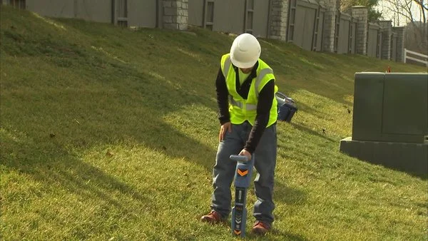 Technician in a hard hat and safety vest using a utility locator on a grassy slope near underground vaults.