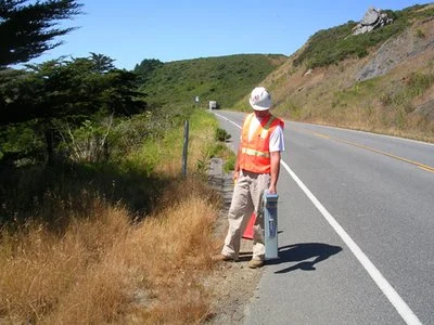Technician in an orange safety vest locating utilities along a hillside road.