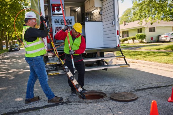 Two technicians in safety gear lowering a mainline inspection crawler into a manhole from an equipment truck.