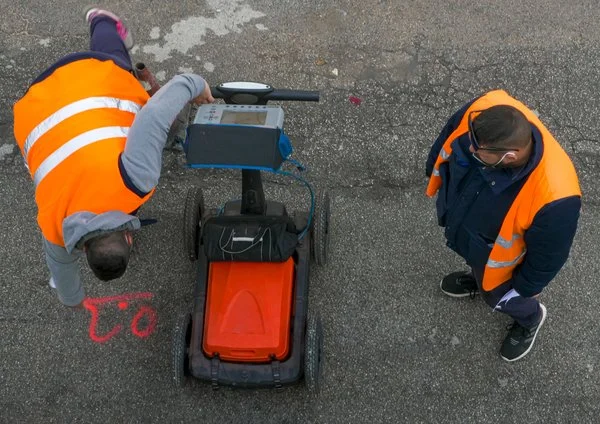Two technicians in safety vests operating a ground penetrating radar cart while surveying a paved street from an overhead view.