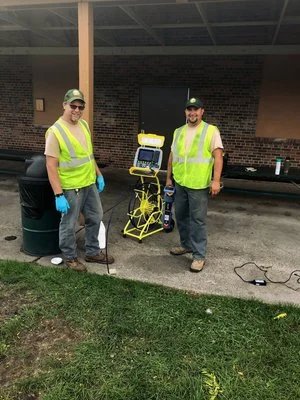 Two technicians in high-visibility vests standing with a yellow locating unit under a shelter.